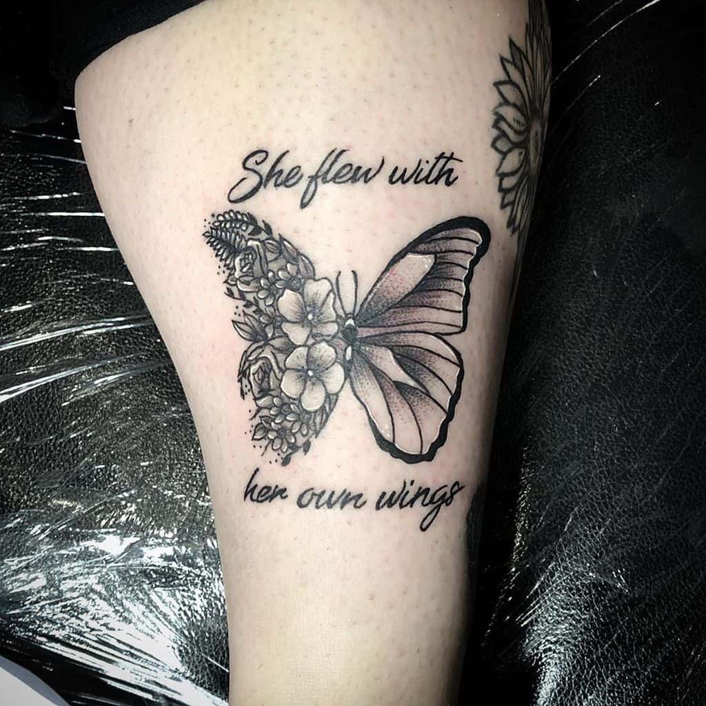 Elegant black and grey butterfly and floral tattoo with the quote 'She flew with her own wings,' symbolizing strength and independence.