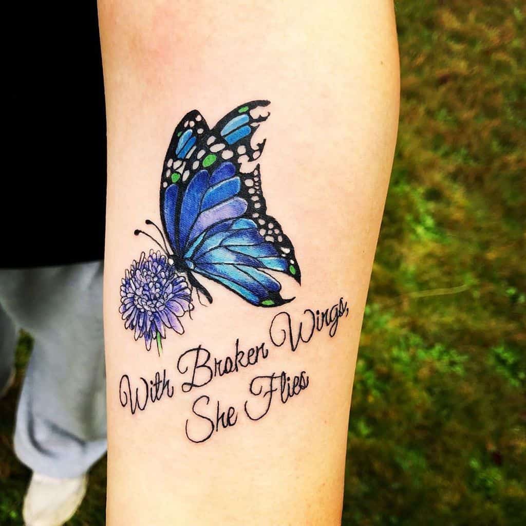 Vibrant blue butterfly tattoo with floral details and the quote 'With Broken Wings, She Flies,' symbolizing resilience and strength.