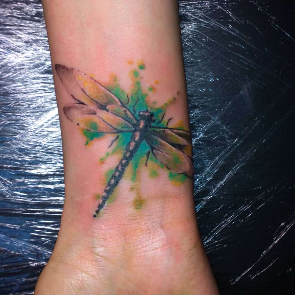Vibrant watercolor dragonfly tattoo on the wrist, featuring splashes of green and yellow for a dynamic and artistic effect.