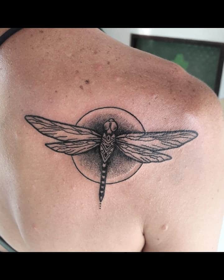 Black ink dragonfly tattoo on the shoulder with geometric detailing and a circular background. The intricate design adds a bold effect.