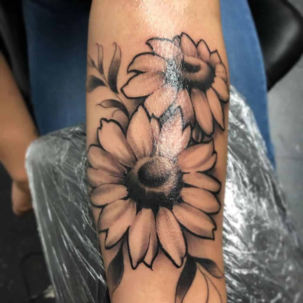 Bold black and grey sunflower tattoo on forearm with soft shading and intricate petal details, featuring two large flowers and leaves.