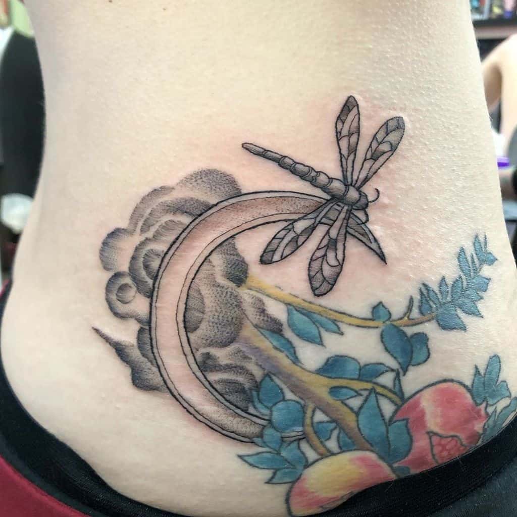 Detailed dragonfly tattoo with a crescent moon, clouds, and colorful floral elements, placed on the side of the torso.