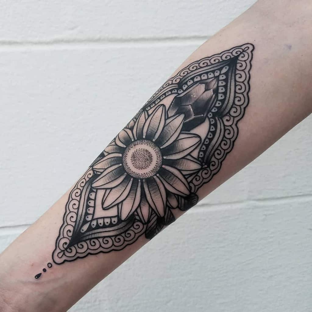 Black and grey forearm tattoo featuring a detailed sunflower surrounded by an ornate mandala-style frame with intricate patterns.