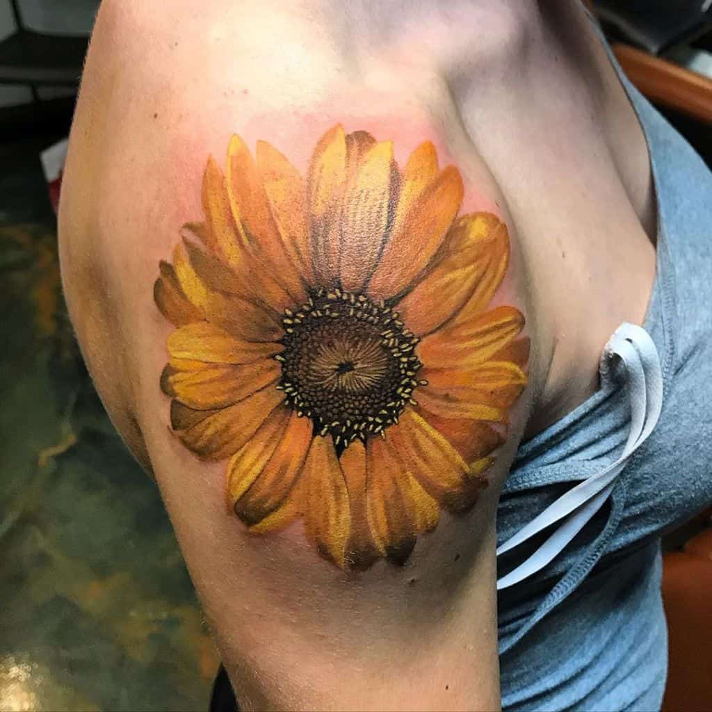Large, realistic sunflower tattoo on upper arm with vibrant yellow and orange petals, a detailed dark center, and smooth shading.