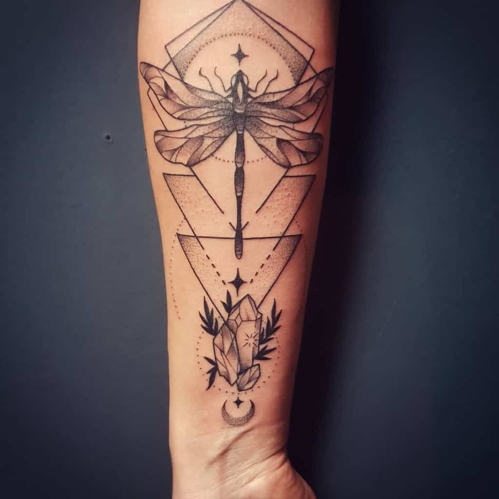 Geometric dragonfly tattoo on the forearm, featuring fine linework, dot shading, crystals, and celestial elements for a mystical design.