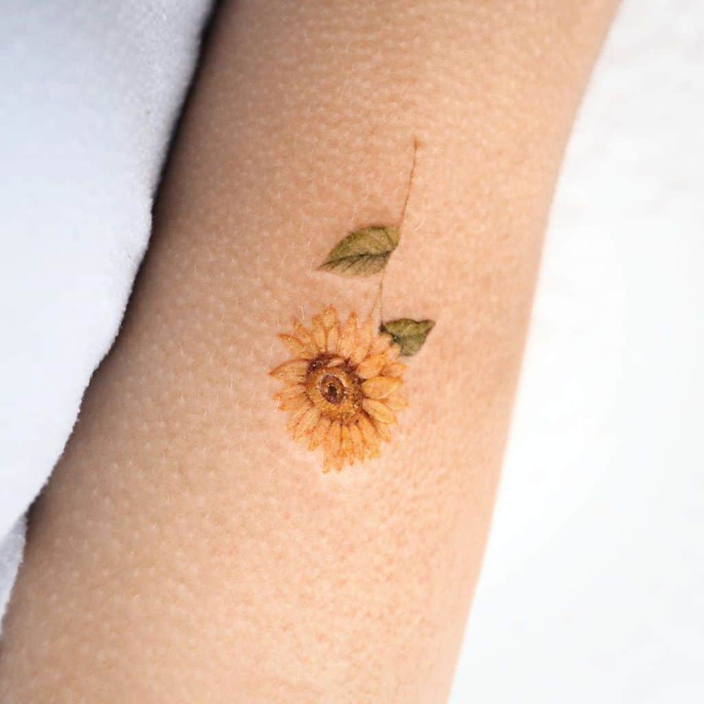 Tiny, delicate sunflower tattoo on forearm with fine details, soft yellow petals, a thin stem, and small green leaves for a subtle design.