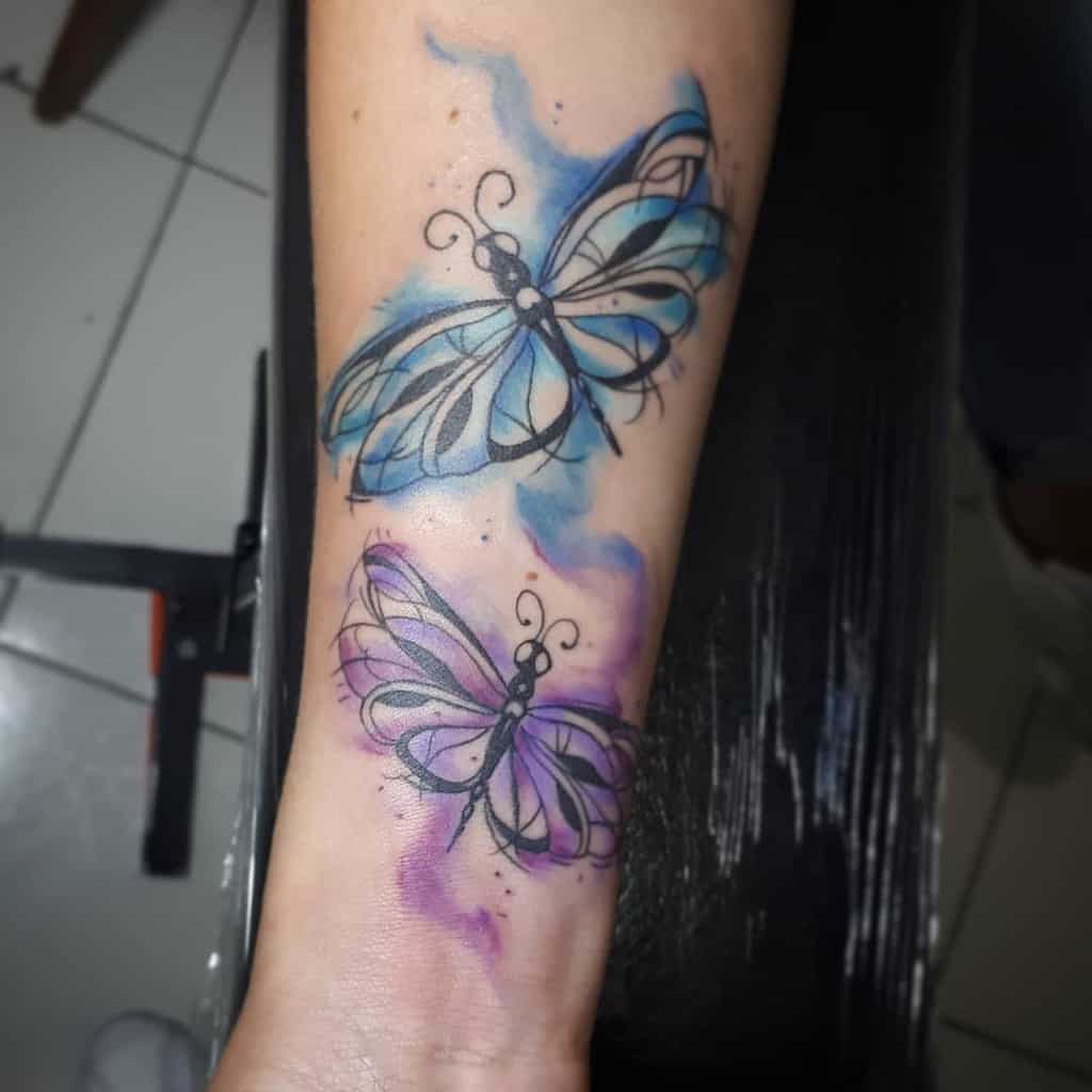 Artistic watercolor dragonfly tattoo on the forearm, featuring two stylized designs with flowing blue and purple accents for a dreamy effect.