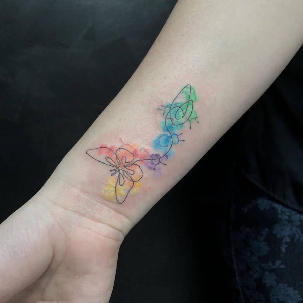 Minimalist butterfly tattoo on the wrist with fine black outlines and a soft rainbow watercolor background for a delicate, artistic look.