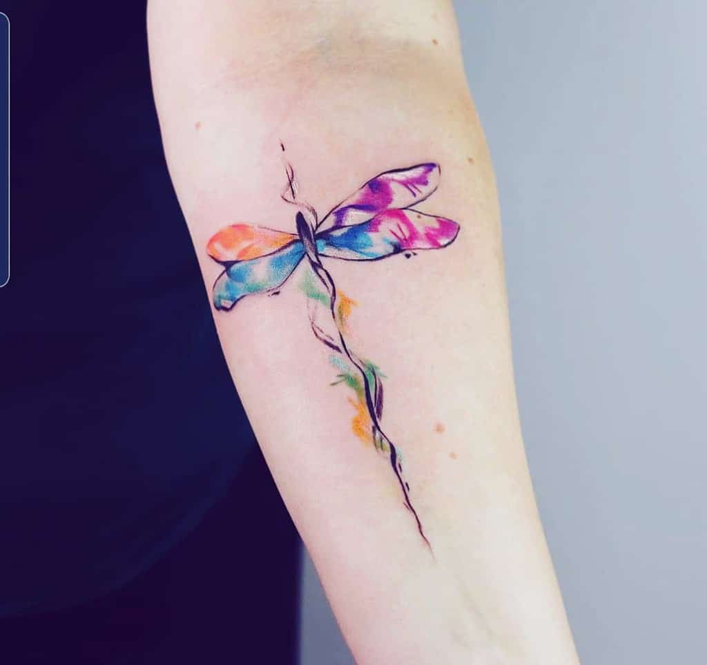Vibrant watercolor dragonfly tattoo on the forearm, featuring multicolored wings and a delicate, artistic stem-like body design.