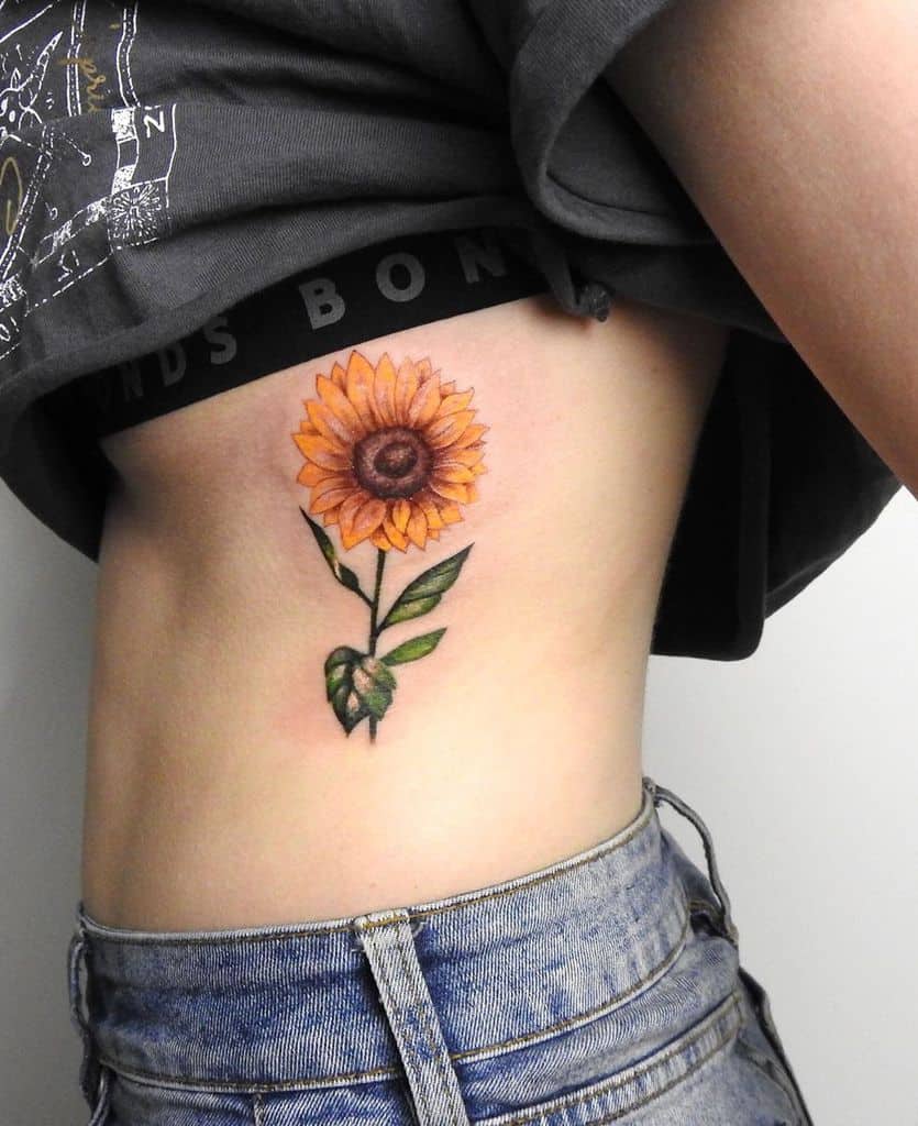 Vibrant sunflower tattoo on ribcage with detailed yellow petals, a brown center, and green leaves, creating a bold and realistic floral design.