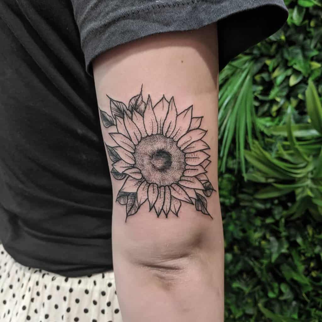 Black ink sunflower tattoo on upper arm with fine line details and shading, featuring bold petals and a textured center.