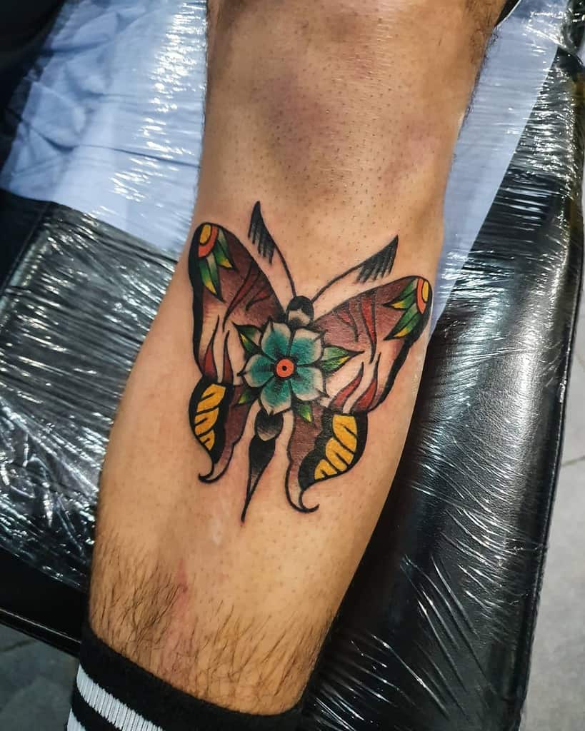 Traditional-style butterfly tattoo on the leg, featuring earthy tones, bold black outlines, and a vibrant blue flower at the center.