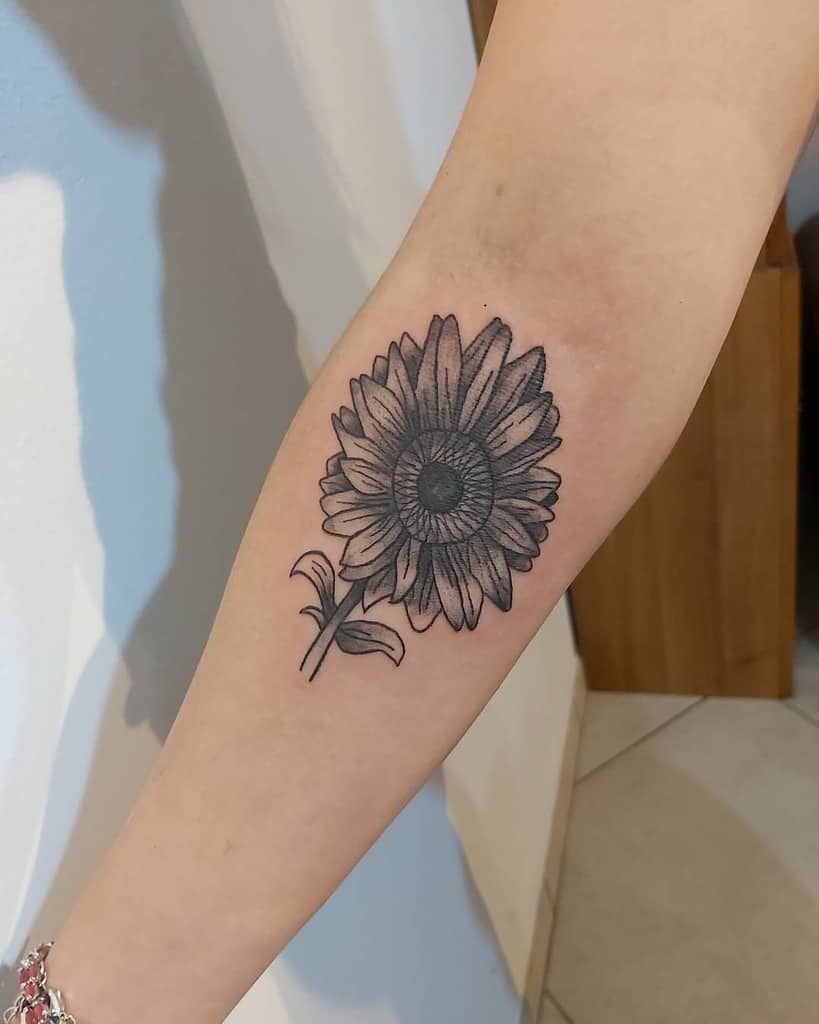Black and grey sunflower tattoo on forearm with fine line details and shading, featuring bold petals and a delicate stem.