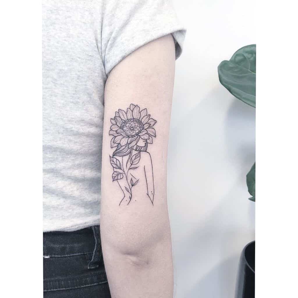 Minimalist black ink tattoo on upper arm featuring a sunflower growing from an abstract human figure, with fine line details and a delicate design.