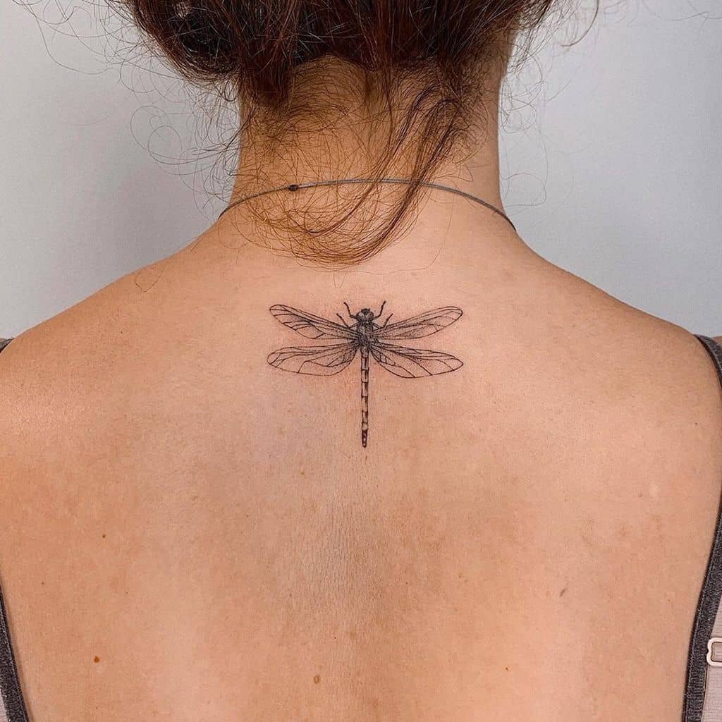 Minimalist dragonfly tattoo in fine black ink, placed at the upper back between the shoulders, featuring delicate wing details.