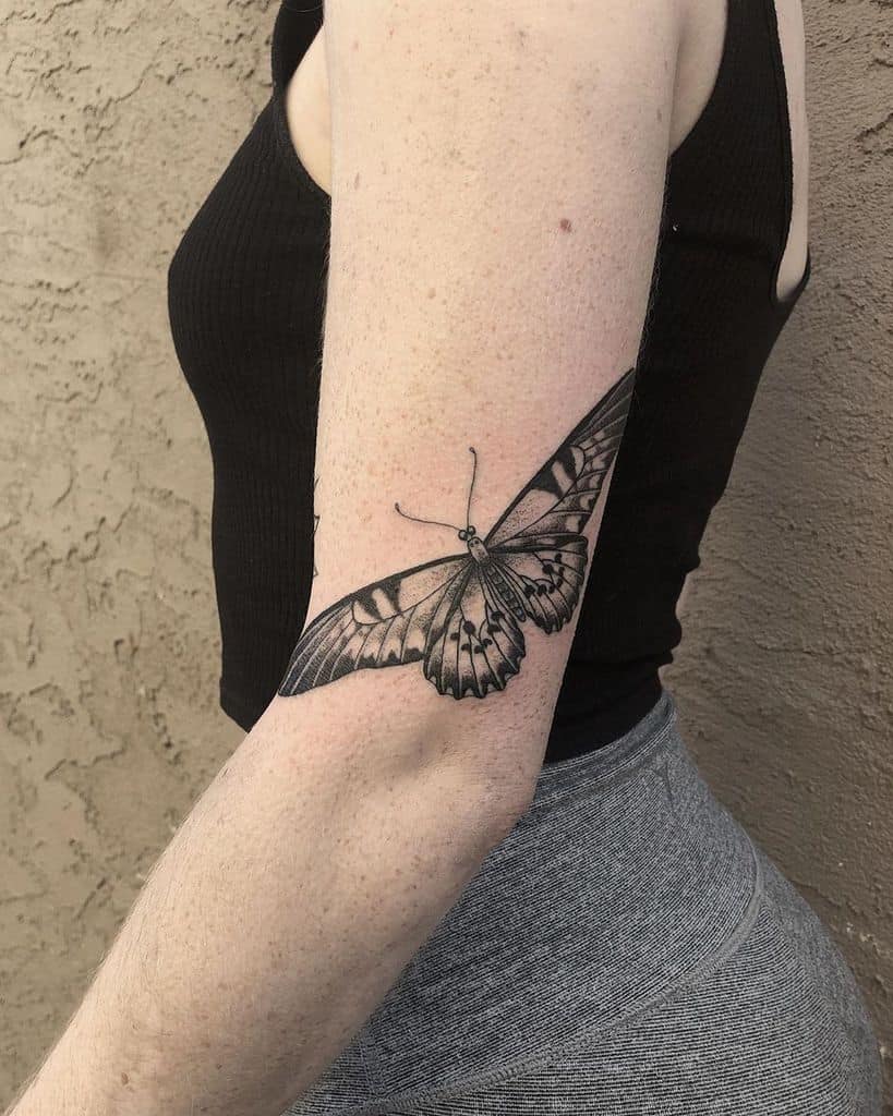 Black and gray butterfly tattoo wrapping around the arm, featuring intricate shading and fine details for a bold yet delicate design.