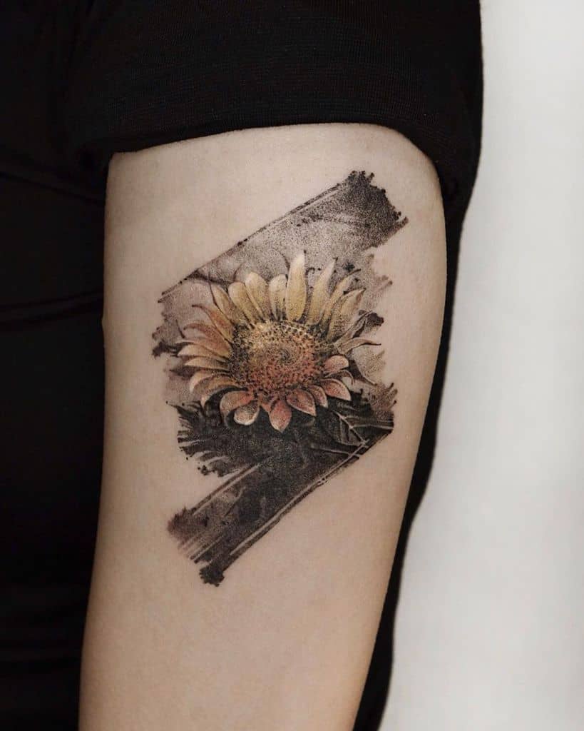 Realistic sunflower tattoo on upper arm with a textured, brushstroke-style background, featuring fine details and warm yellow tones.