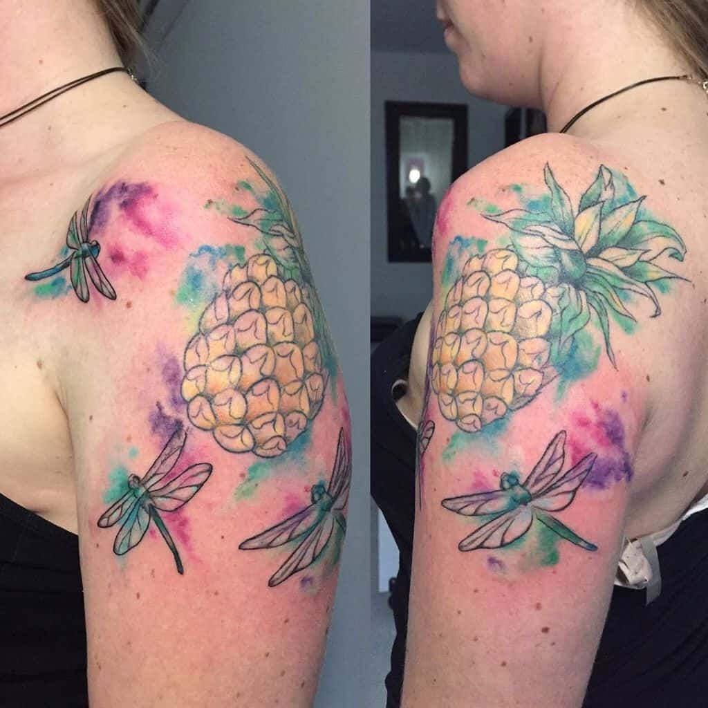 Vibrant tropical-themed tattoo on the upper arm featuring dragonflies, pineapples, and watercolor splashes in pink, blue, and green.