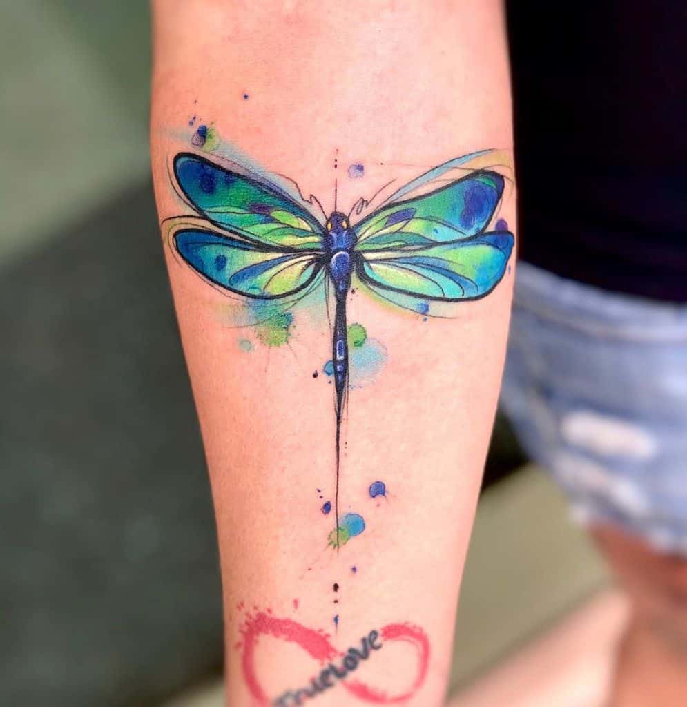 Vibrant watercolor dragonfly tattoo on the forearm, featuring bold blue and green hues with artistic splashes for a dynamic effect.