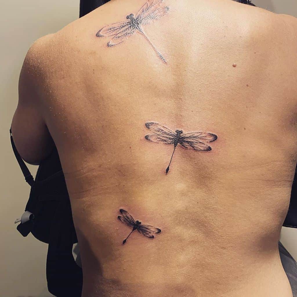 Three delicate dragonfly tattoos in black ink, arranged diagonally across the back. The fine shading adds a soft, natural effect.