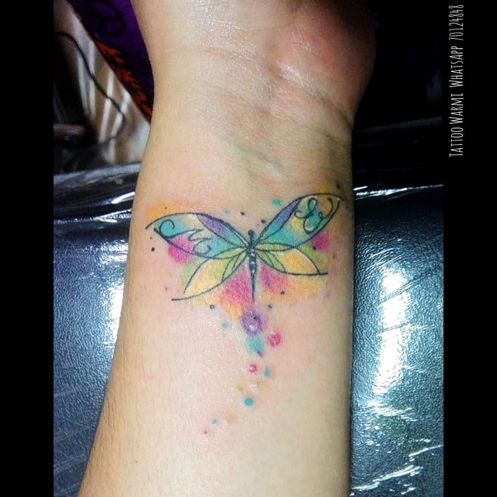 Colorful watercolor dragonfly tattoo on the wrist, featuring delicate swirls and vibrant splashes of blue, pink, yellow, and purple.