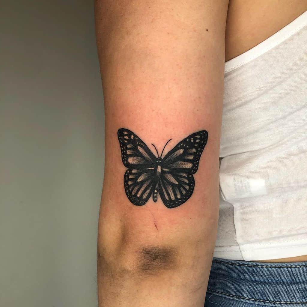 Black butterfly tattoo on the upper forearm, featuring bold shading and intricate wing details for a striking and elegant design.