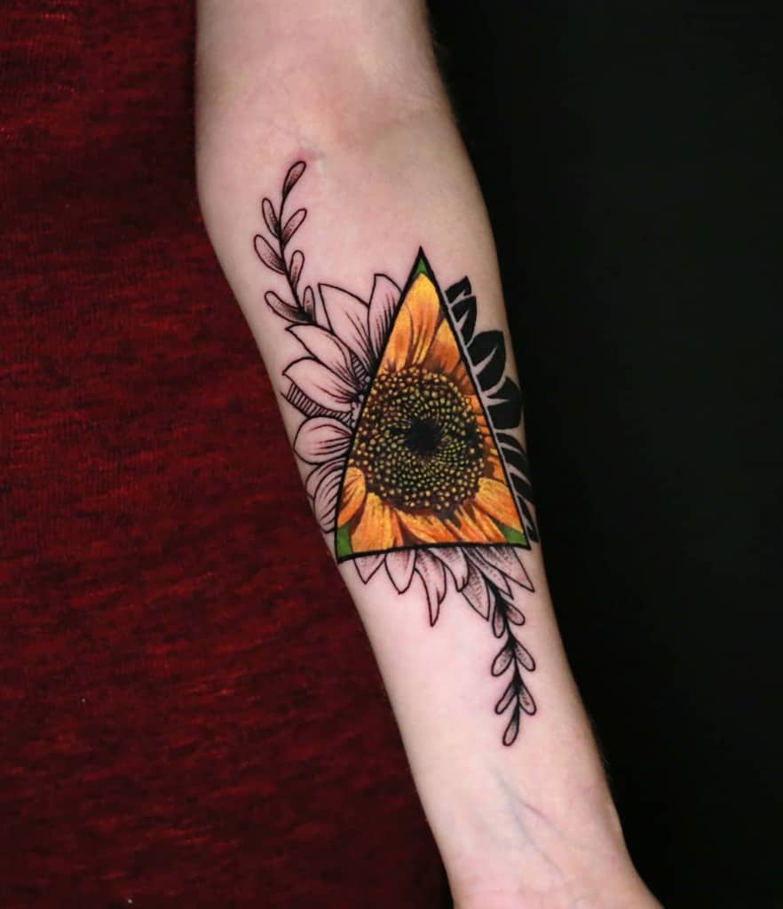 Forearm tattoo featuring a vibrant sunflower inside a geometric triangle, with black and grey floral accents extending beyond the shape.