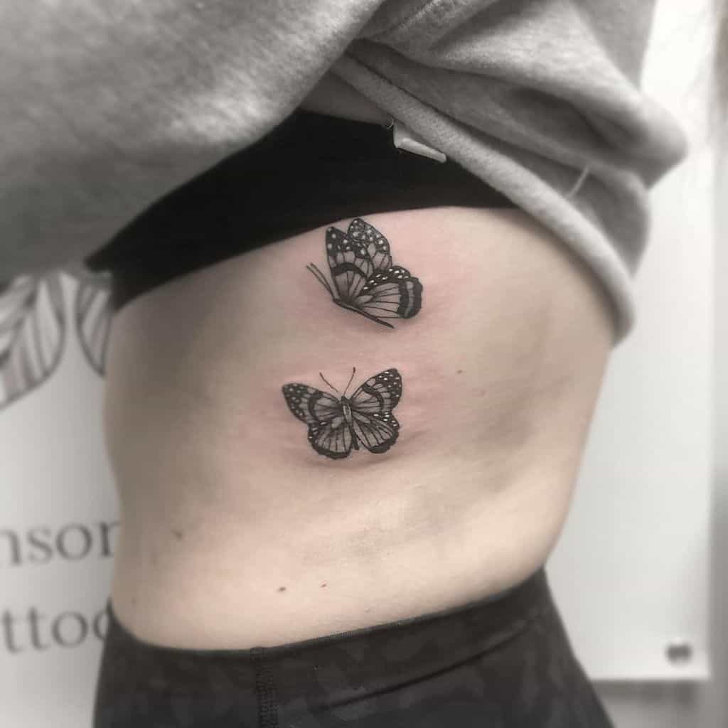 Two black and gray butterfly tattoos on the ribcage, featuring fine shading and delicate wing details for a soft, elegant design.