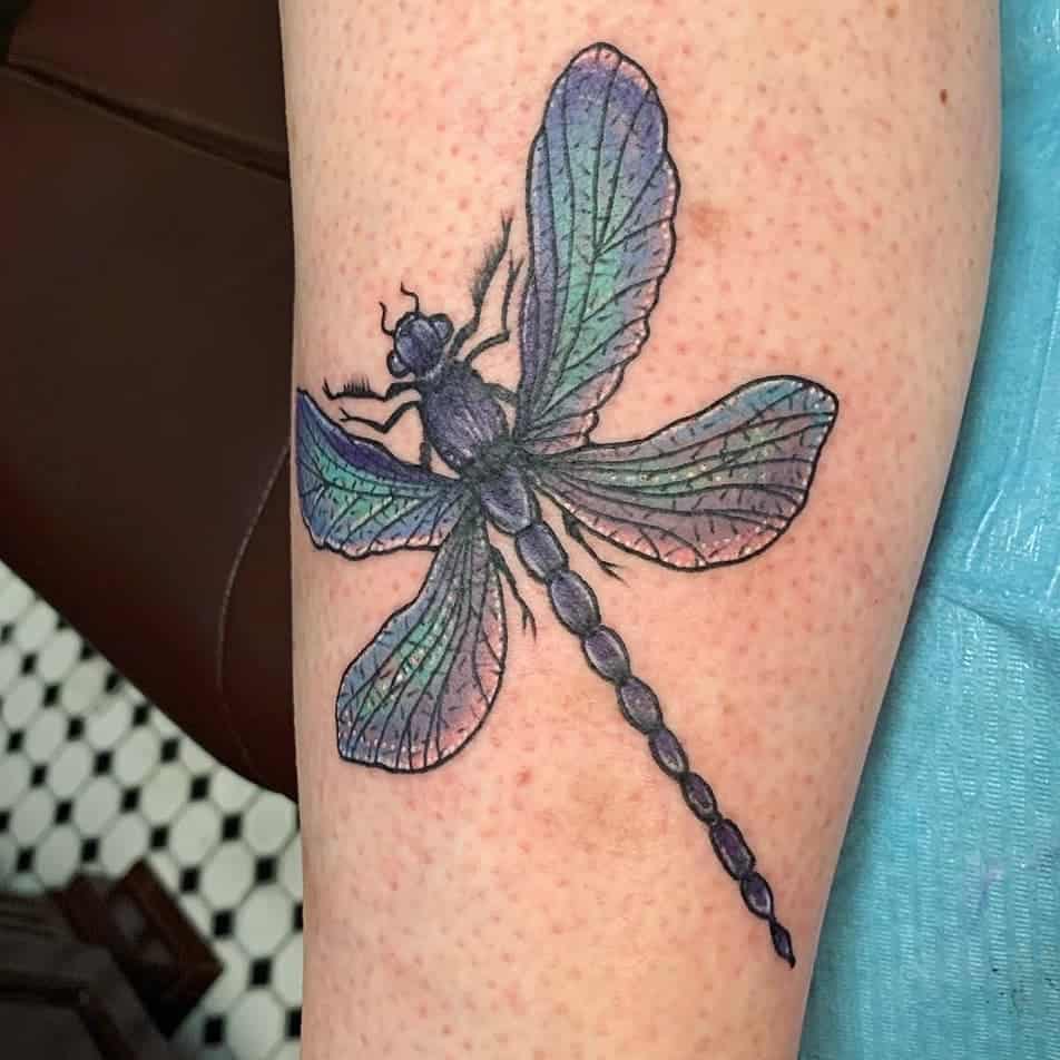 Realistic dragonfly tattoo on the leg with iridescent blue and green wings, fine details, and bold black outlines.