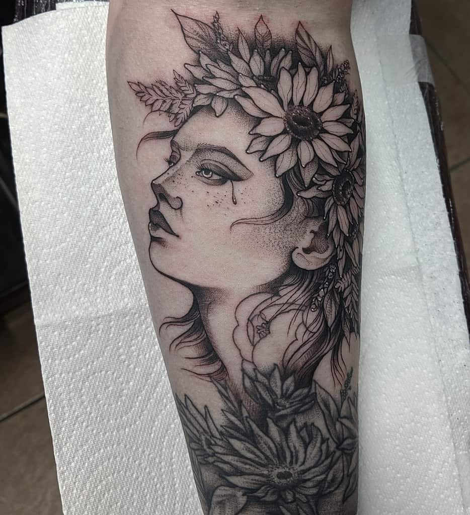 Black and grey forearm tattoo of a woman's face with flowing hair, adorned with a sunflower crown, featuring fine line shading and intricate details.