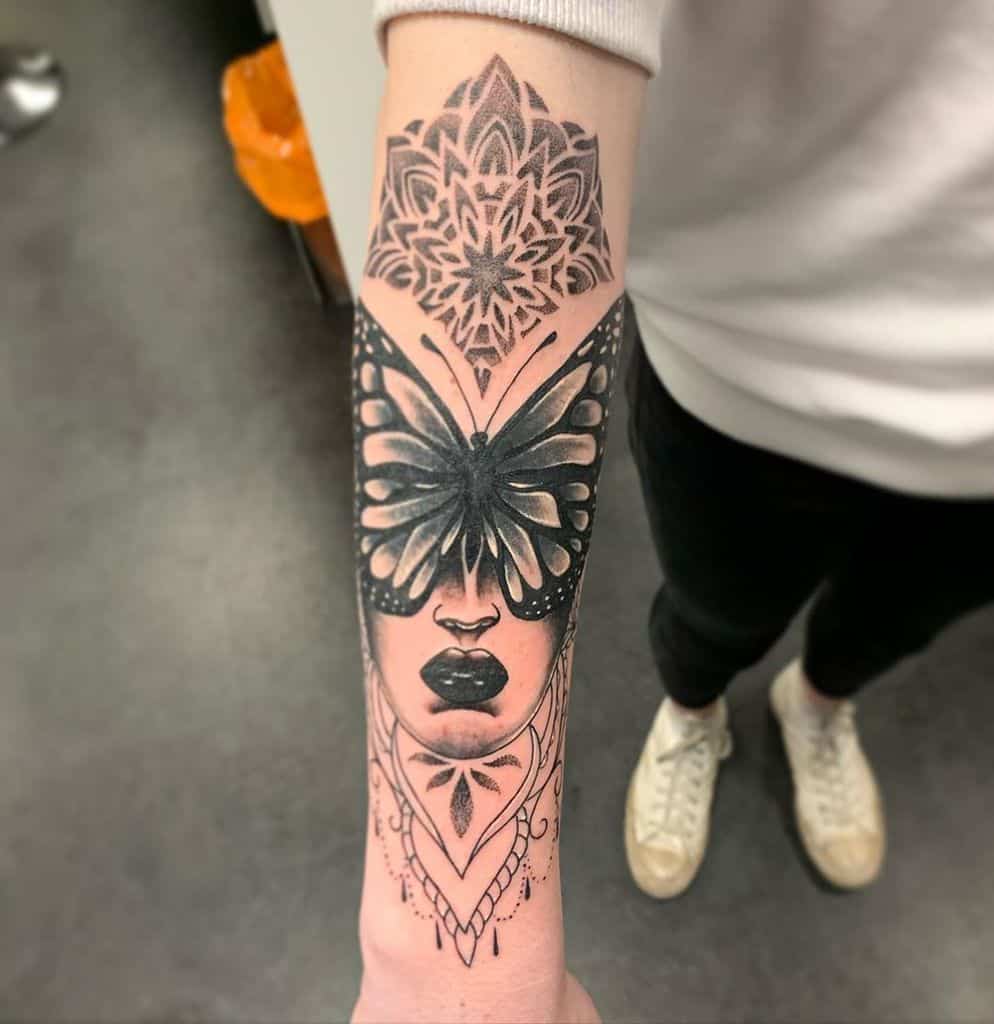 Intricate black and grey tattoo of a woman's face with a butterfly mask and mandala design, blending surrealism with geometric elements.