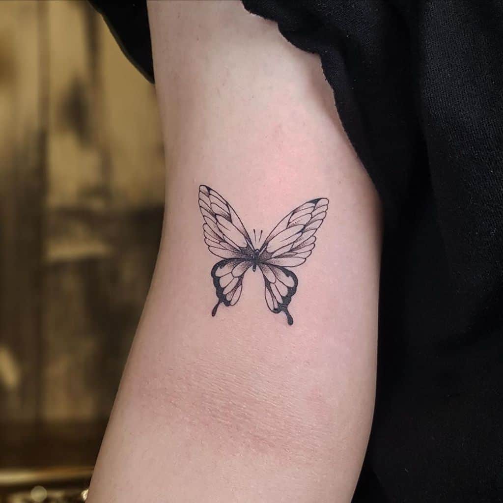 Minimalist black and grey butterfly tattoo on the upper arm, symbolizing transformation, freedom, and elegance.