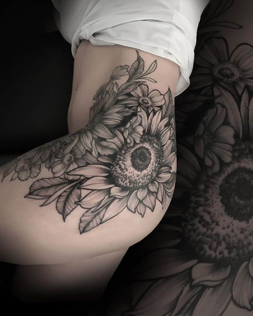 Large black and grey tattoo covering hip and thigh, featuring a detailed sunflower with intricate shading, surrounding flowers, and leaves.