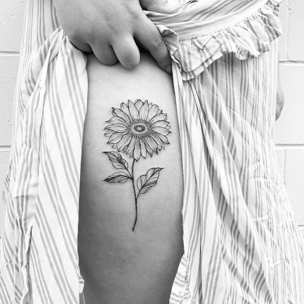 Black and grey sunflower tattoo on thigh with fine line details, delicate shading, and a long, thin stem for a minimalist floral design.