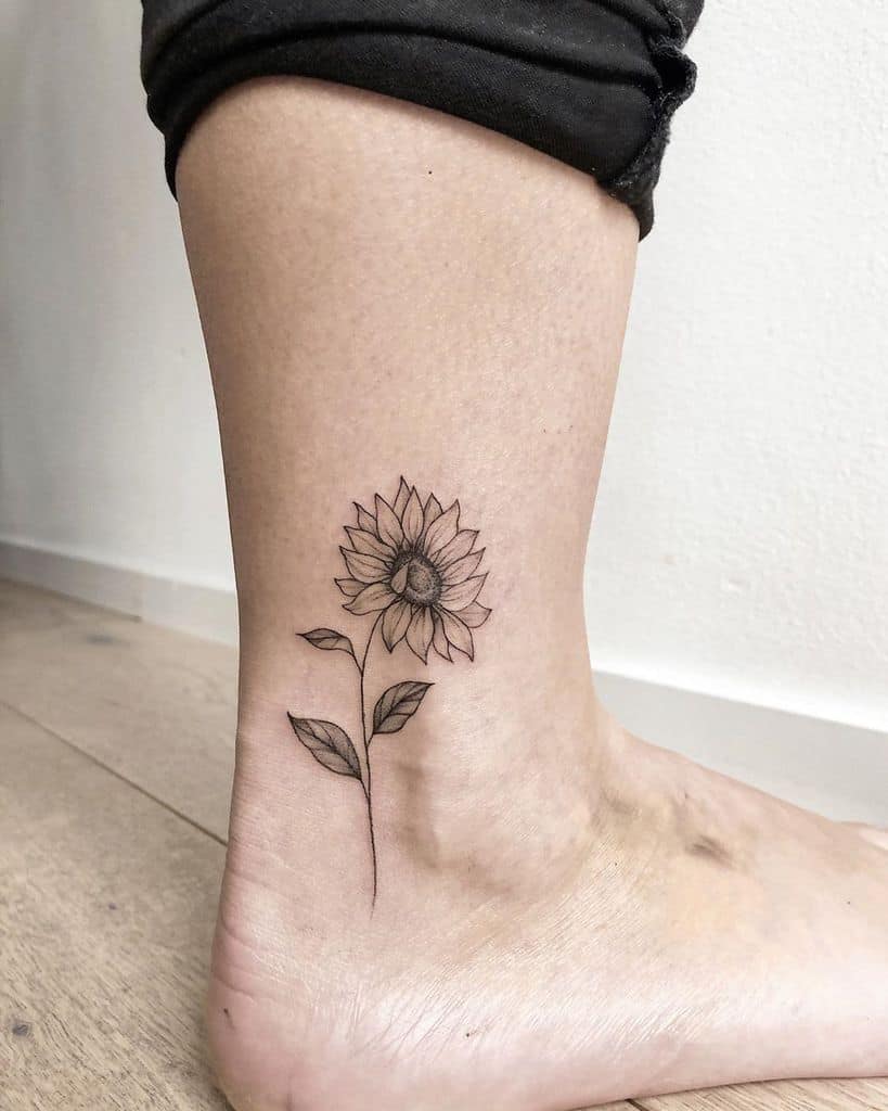 Minimalist black ink sunflower tattoo on the ankle with fine line details, a thin stem, and delicate leaves for a subtle floral design.