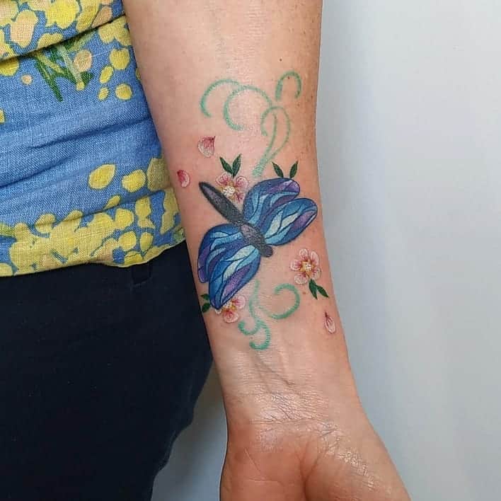 Colorful dragonfly tattoo on the wrist with blue wings, floral details, and swirling green accents for a delicate, nature-inspired look.