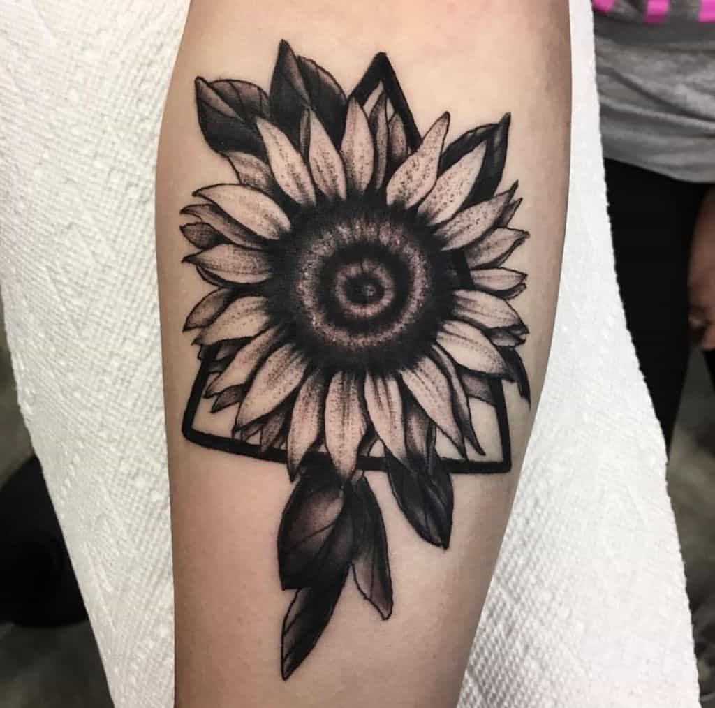 Bold black and grey sunflower tattoo on forearm with geometric triangle framing, deep shading, and intricate petal and leaf details.