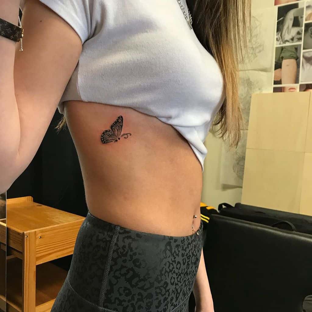 Minimalist black butterfly tattoo on the ribcage, adding a delicate and feminine touch to the design.