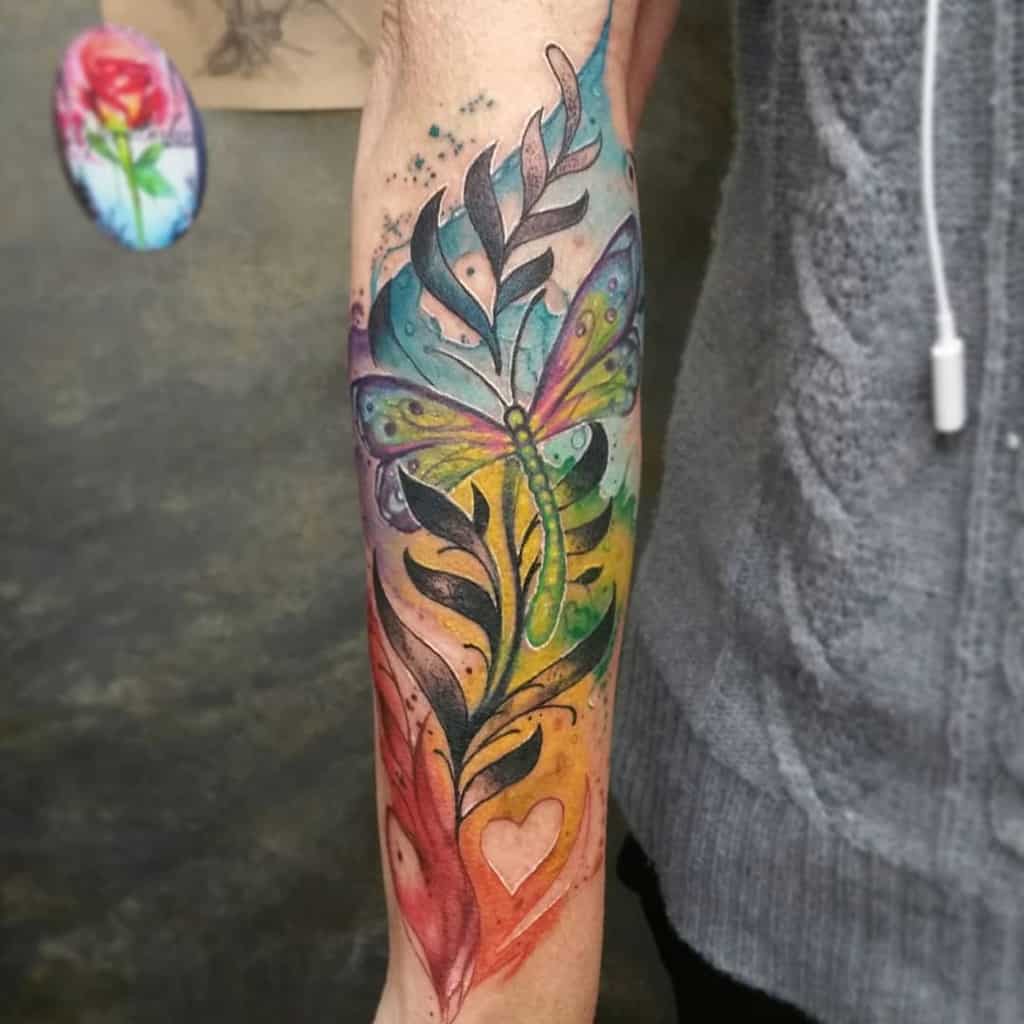 Vibrant watercolor dragonfly tattoo on the forearm, featuring rainbow hues, botanical elements, and artistic abstract details.