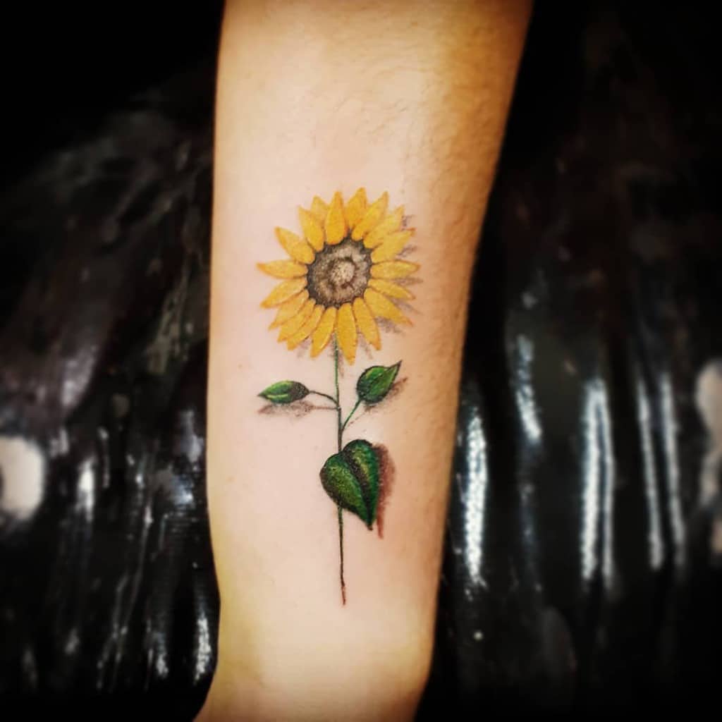 Realistic sunflower tattoo on forearm with bright yellow petals, a brown textured center, and green leaves, featuring soft shading for depth.