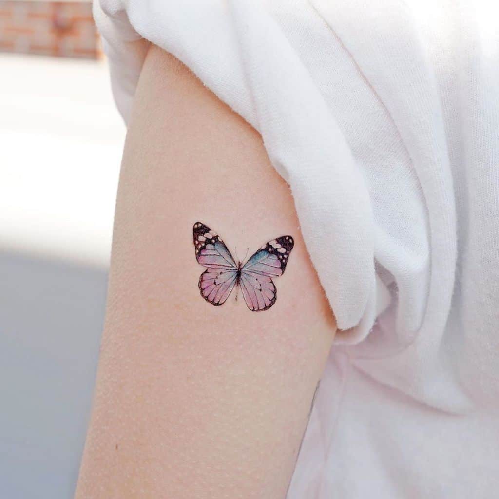 Minimalist pastel butterfly tattoo on the upper arm with soft pink and blue hues, symbolizing transformation and beauty.