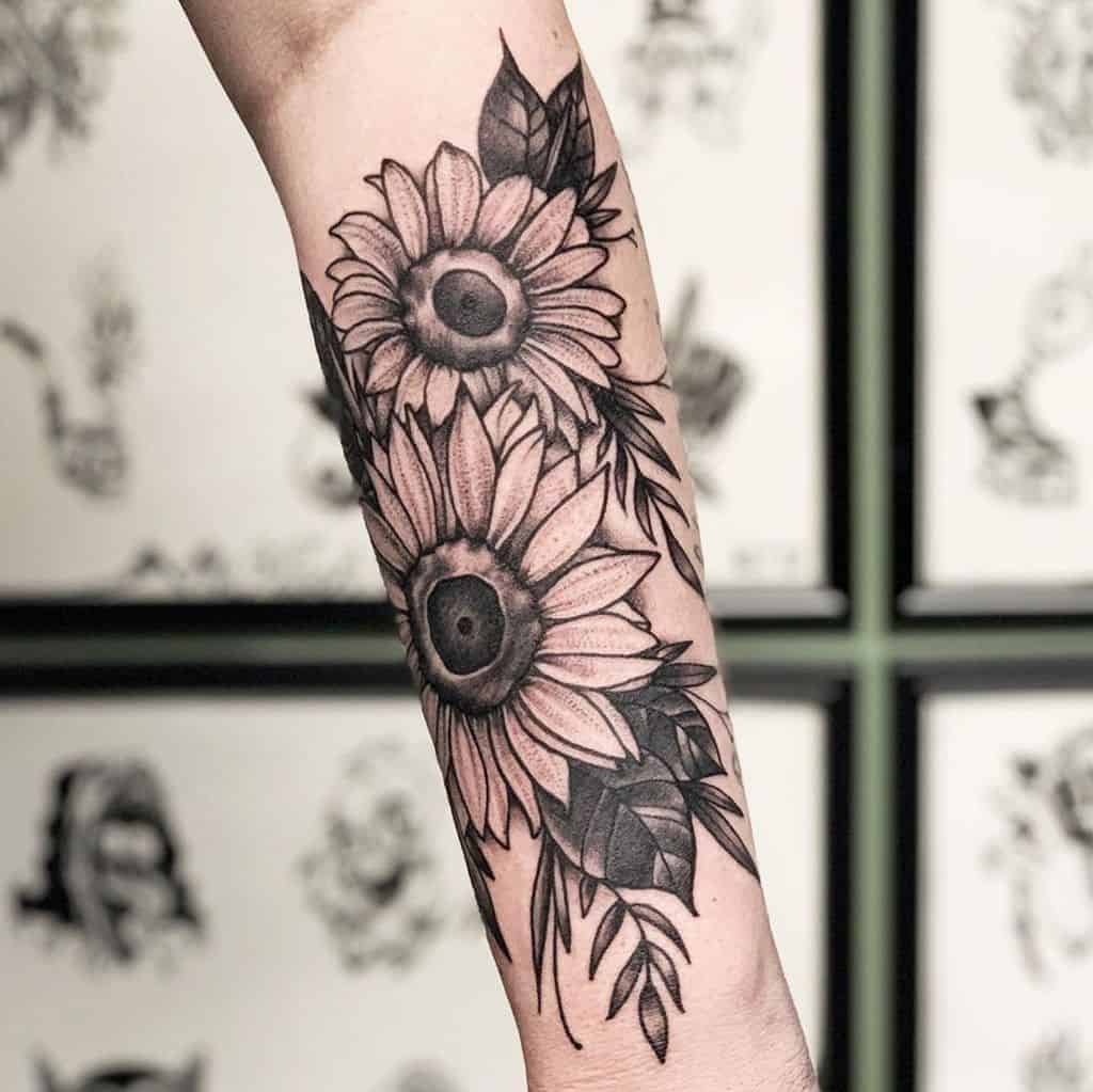 Black and grey forearm tattoo featuring two detailed sunflowers with bold shading, fine line leaves, and intricate petal details.