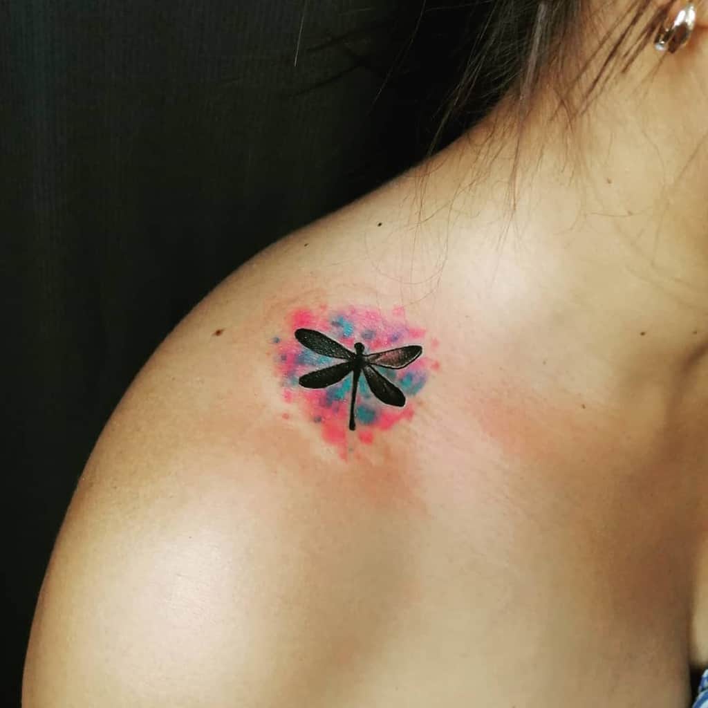 Black dragonfly tattoo on the shoulder with a vibrant watercolor background in red and blue hues for a striking effect.