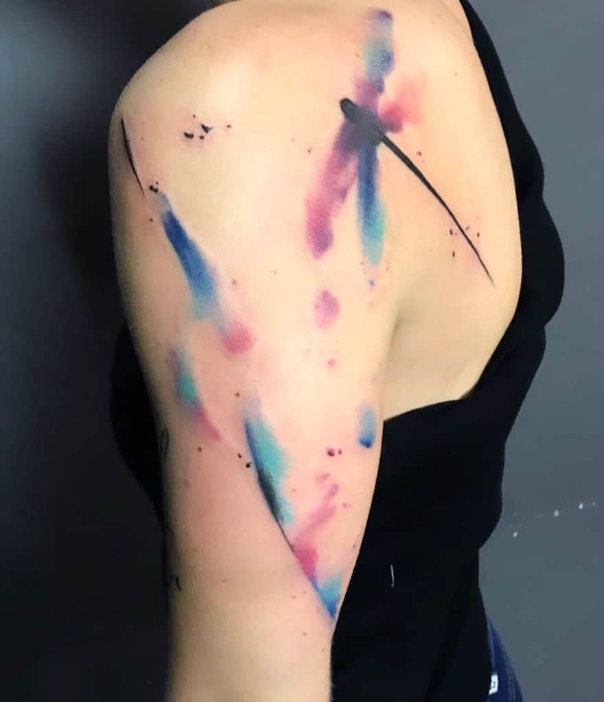 Abstract watercolor dragonfly tattoo on the shoulder and upper arm, featuring bold splashes of blue, pink, and black ink for a fluid, artistic look.
