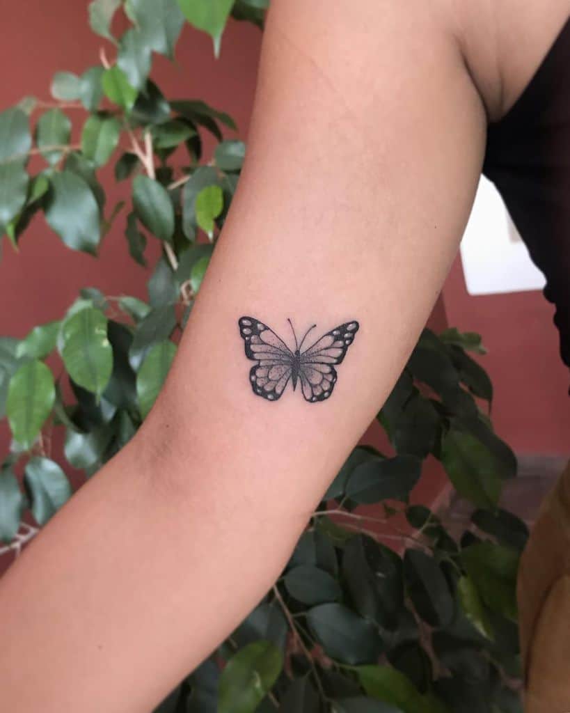 Small black and gray butterfly tattoo on the upper arm, featuring fine dot shading and delicate wing details for a subtle, elegant look.
