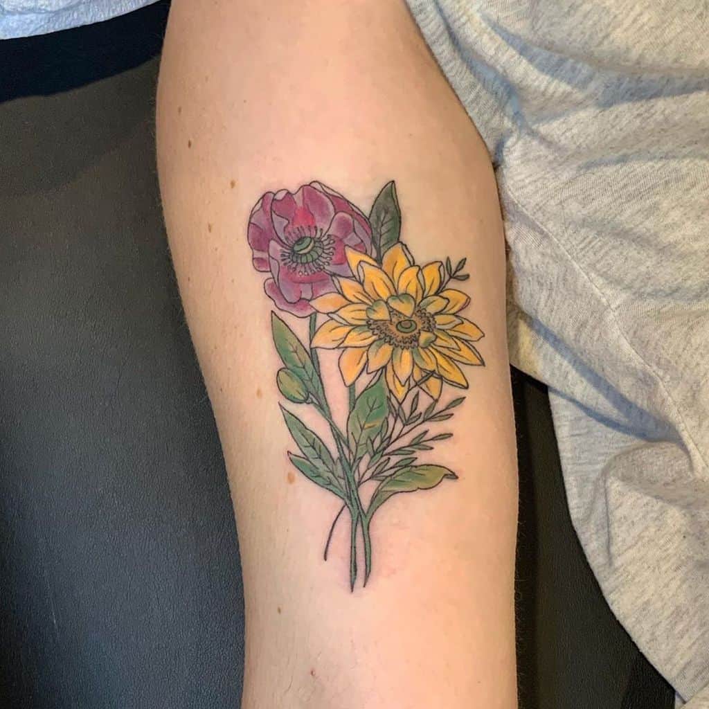 Colorful floral tattoo on upper arm featuring a bright yellow sunflower and a pink poppy, with green leaves and fine line details.