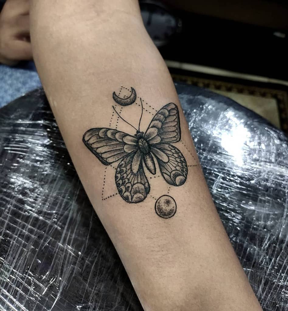 Black and gray butterfly tattoo on the forearm, featuring intricate dotwork, geometric shapes, a crescent moon, and a celestial theme.