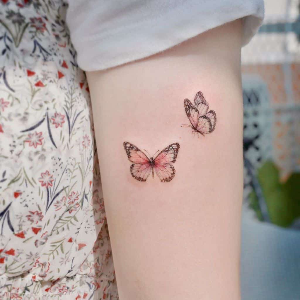 Delicate pink butterfly tattoo design with two butterflies on the upper arm, symbolizing grace, transformation, and freedom.