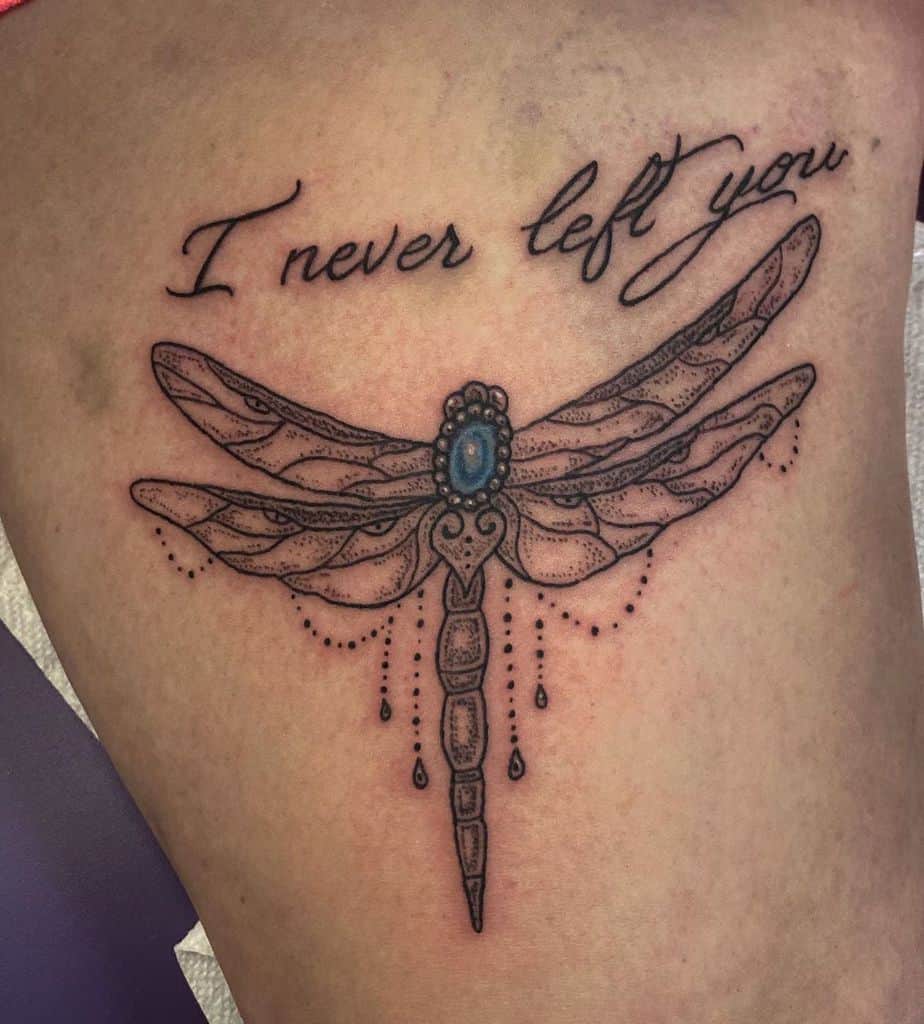 Ornamental dragonfly tattoo with a blue gemstone and delicate bead details, paired with cursive script reading 'I never left you.'