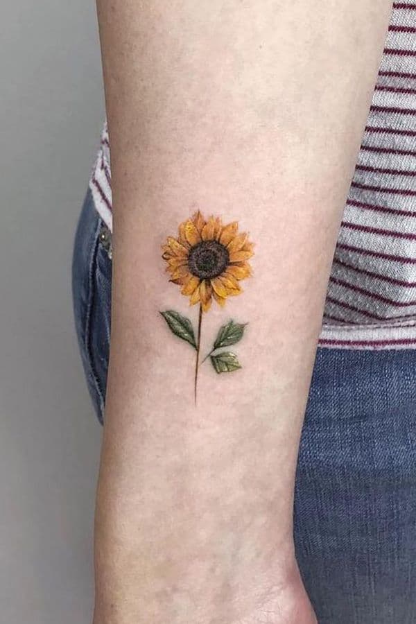 Small, realistic sunflower tattoo on forearm with fine details, warm yellow petals, a brown center, and green leaves for a delicate floral look.